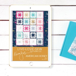 Op de afbeelding: Een digitale tablet toont een quiltpatroon getiteld "American Honey" van Keera Job. Het quiltontwerp heeft sterblokken in roze, blauwgroen en crème op een marineblauwe achtergrond. De tablet staat naast een blauw boek.