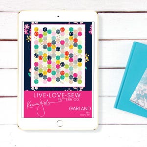 May include: A digital tablet shows a quilt pattern with a grid of colorful circles on a dark blue background. The tablet screen displays the text "LIVE.LOVE.SEW" and "GARLAND" with dimensions of 148.6 cm x 182.9 cm. A turquoise book is visible.