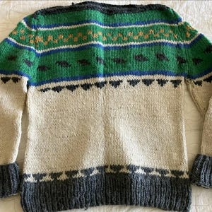 May include: A long-sleeve knit sweater featuring a cream-colored body with a dark gray ribbed hem and cuffs. The upper section displays horizontal bands of green, blue, and orange patterns, including small dark gray shapes across the chest area.