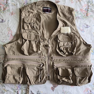 May include: Tan utility vest with multiple pockets and a zipper closure. The vest has a rectangular patch with the text "WEATHER RITE" on the upper back. The vest is designed for outdoor activities and has a functional, practical design.