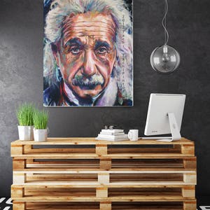 May include: A colorful portrait of Albert Einstein with text on the side. The artwork is displayed above a wooden pallet-style cabinet. The cabinet holds a computer monitor, books, a mug, and small potted plants.