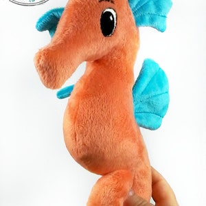 Peut inclure: Un hippocampe en peluche orange avec des nageoires bleues. L'hippocampe a de grands yeux noirs et un sourire cousu. L'hippocampe est tenu par une main.