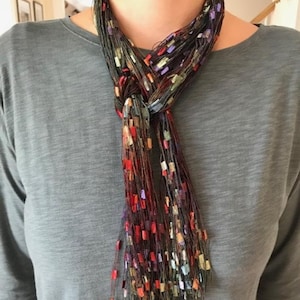 May include: A knotted scarf with a dark base and colorful rectangular accents in red, orange, purple, and green. The scarf is draped over a gray shirt, creating a contrast of textures and colors.