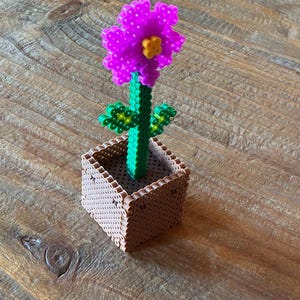 Peut inclure: Une fleur artisanale faite de perles en plastique, avec une tête de fleur violette vibrante et un centre jaune, une tige et des feuilles vertes. La fleur est dans un pot carré marron, le tout sur une surface en bois.