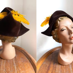 Può includere: Un cappello marrone vintage con una tesa larga e un gruppo decorativo di piume giallo brillante. Il cappello è esposto su una testa di manichino, che ne mette in risalto la forma e lo stile. Il colore scuro del cappello contrasta con le piume vivaci.