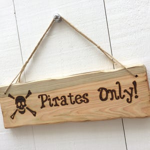 Puede incluir: Un letrero de madera con una calavera y huesos cruzados y el texto "Pirates Only!" colgado de una cuerda.