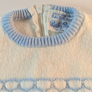 May include: Close-up of a vintage baby sweater. The sweater is off-white with light blue trim around the neck and bottom. A zipper runs down the front. The label reads "Saks Fifth Avenue Baby Boutique."
