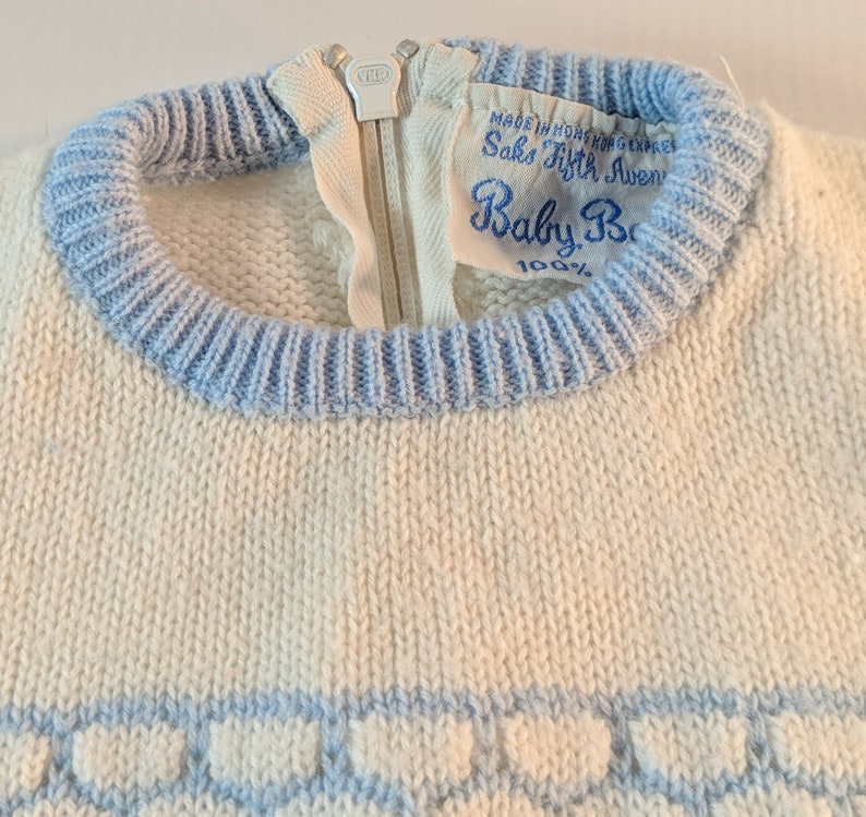 May include: Close-up of a vintage baby sweater. The sweater is off-white with light blue trim around the neck and bottom. A zipper runs down the front. The label reads "Saks Fifth Avenue Baby Boutique."