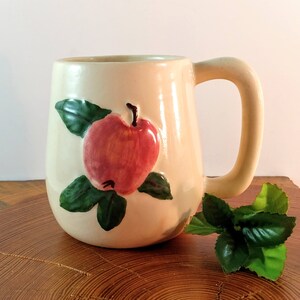 May include: A cream-colored ceramic mug with a red apple and green leaves design. The mug has a rounded body and a handle.