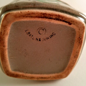May include: A vintage ceramic pitcher with a brown glaze and a green floral design. The bottom of the pitcher has the text "C.Baltine & Sun Inc." in black.