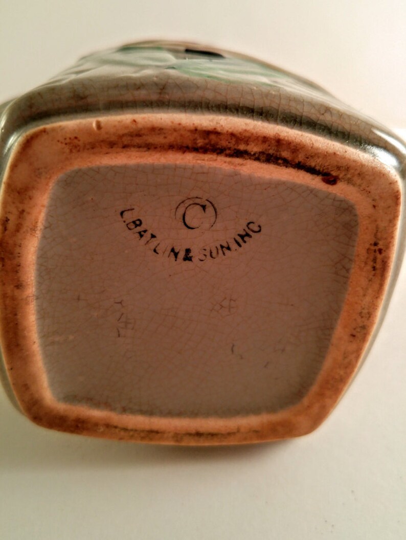 May include: A vintage ceramic pitcher with a brown glaze and a green floral design. The bottom of the pitcher has the text "C.Baltine & Sun Inc." in black.