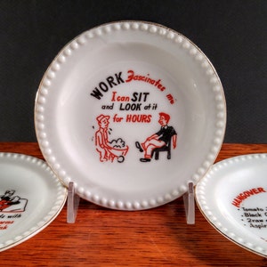 May include: Three white ceramic plates with gold trim and black and red text. The center plate reads "WORK Fascinates me I can SIT and LOOK at it for HOURS". The plate on the left reads "sink And ends with your arms in the sink". The plate on the right reads "HANGOVER BREAKFAST Tomato Juice Black Coffee 2 raw eggs Aspirin".