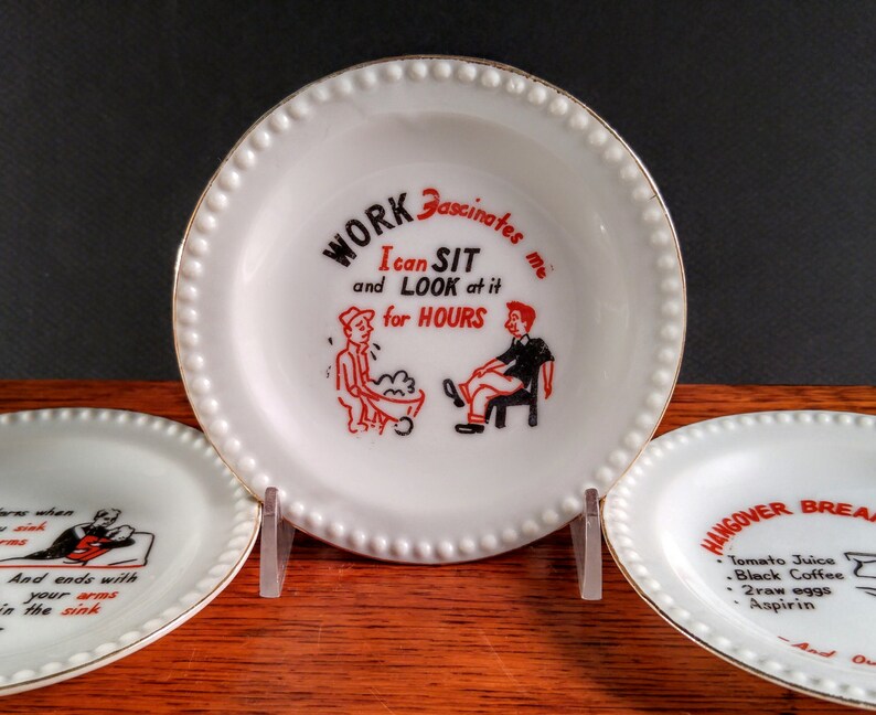 May include: Three white ceramic plates with gold trim and black and red text. The center plate reads "WORK Fascinates me I can SIT and LOOK at it for HOURS". The plate on the left reads "sink And ends with your arms in the sink". The plate on the right reads "HANGOVER BREAKFAST Tomato Juice Black Coffee 2 raw eggs Aspirin".