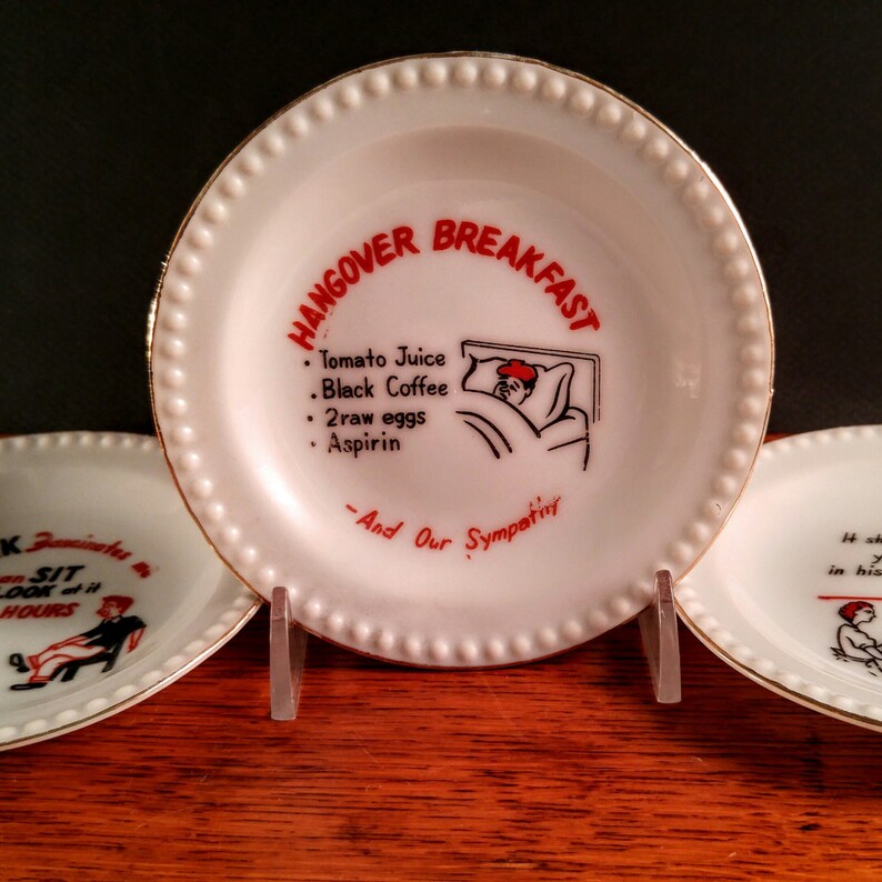 May include: Three white ceramic plates with gold trim. The center plate has a red and black illustration of a person in bed with the text "HANGOVER BREAKFAST" and a list of hangover cure ingredients: "Tomato Juice, Black Coffee, 2raw eggs, Aspirin". The text "And Our Sympatny" is below the list.