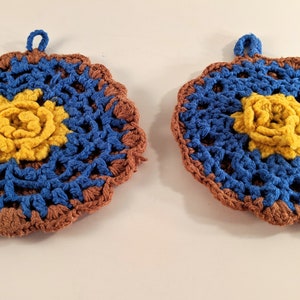 May include: Two crocheted pot holders with a brown, blue, and yellow color scheme. Each pot holder has a flower design in the center.