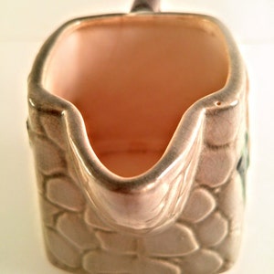 May include: A small, ceramic creamer in the shape of a stone wall. The creamer is glazed in a light brown color with a textured surface that resembles stone. The creamer has a handle and a spout.