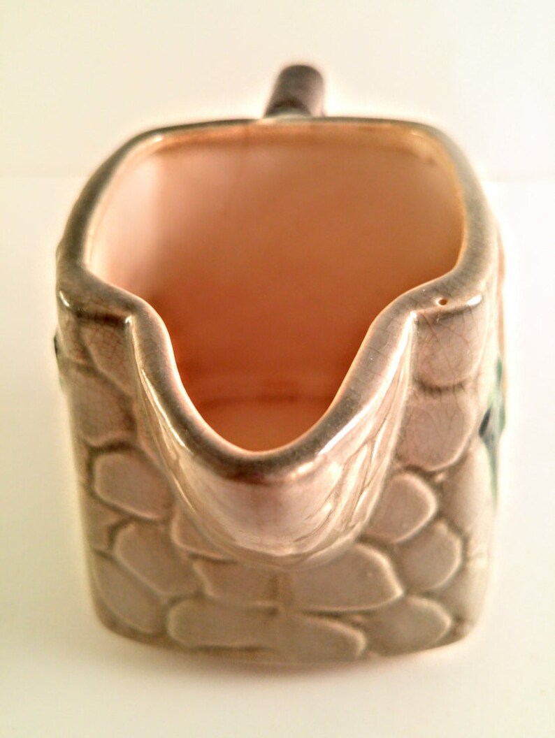 May include: A small, ceramic creamer in the shape of a stone wall. The creamer is glazed in a light brown color with a textured surface that resembles stone. The creamer has a handle and a spout.