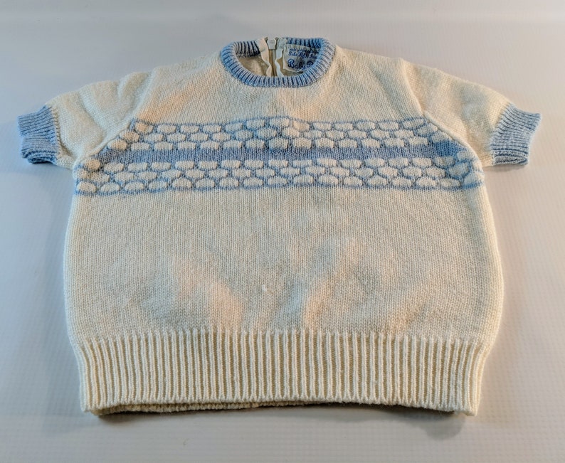 May include: Cream-colored knit short-sleeve sweater with light blue trim on the collar and sleeves. A horizontal band of light blue honeycomb-like pattern is across the chest. The sweater has a ribbed hem and a zipper at the back of the neck.