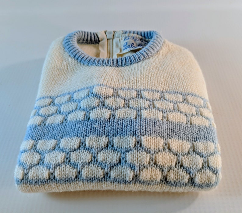May include: Cream-colored knit sweater with a light blue honeycomb pattern across the chest. The sweater has a light blue ribbed collar and a zipper closure at the back. The label reads "Baby".