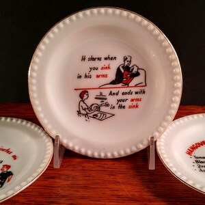 May include: Three white ceramic plates with gold trim and a beaded edge. The center plate has a black and red illustration of a couple with the text "It starts when you sink in his arms and ends with your arms in the sink". The plate on the left has the text "ascinates me T at it S". The plate on the right has the text "HANGOVER Tomato J Black C 2 raw e Aspiri".