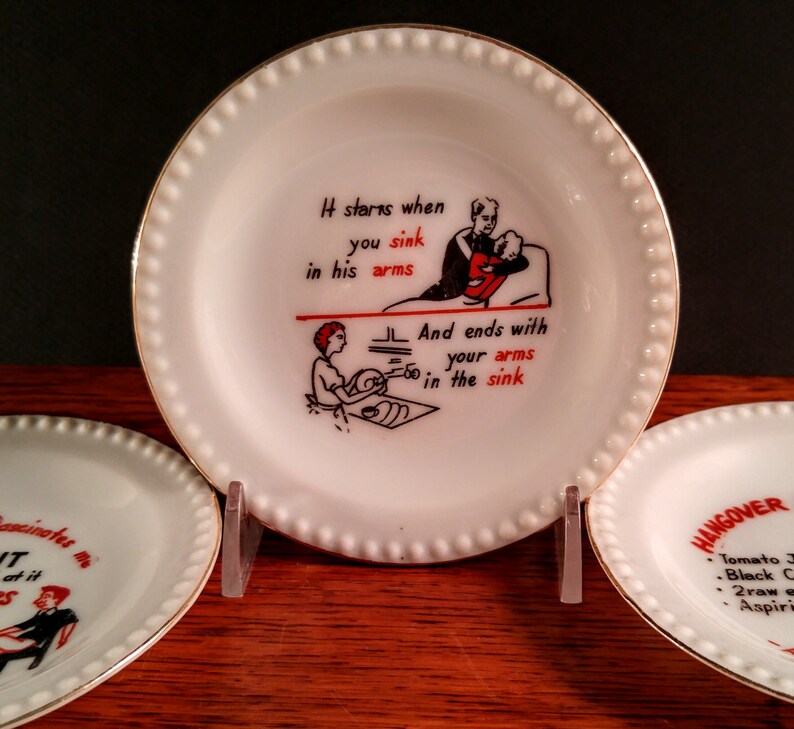 May include: Three white ceramic plates with gold trim and a beaded edge. The center plate has a black and red illustration of a couple with the text "It starts when you sink in his arms and ends with your arms in the sink". The plate on the left has the text "ascinates me T at it S". The plate on the right has the text "HANGOVER Tomato J Black C 2 raw e Aspiri".