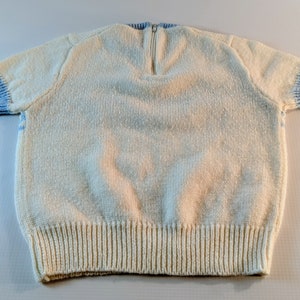 May include: Cream-colored knit sweater with short sleeves and a zipper at the back of the neck. The sleeves and neckline have light blue ribbed trim. The bottom hem is also ribbed. A cozy, handmade garment.