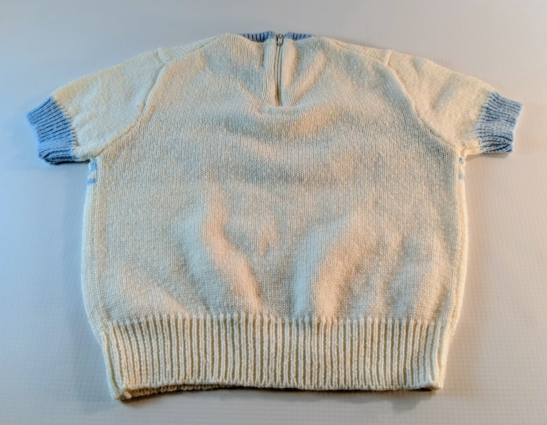 May include: Cream-colored knit sweater with short sleeves and a zipper at the back of the neck. The sleeves and neckline have light blue ribbed trim. The bottom hem is also ribbed. A cozy, handmade garment.
