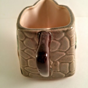 May include: A brown ceramic gravy boat with a stone-like pattern and a brown handle. The boat is shaped like a house and has a light pink interior.