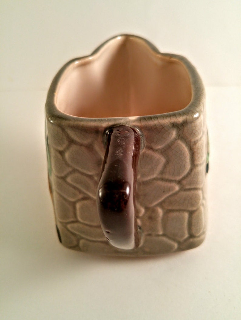 May include: A brown ceramic gravy boat with a stone-like pattern and a brown handle. The boat is shaped like a house and has a light pink interior.