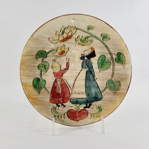 May include: Round ceramic wall hanging featuring a painted scene of two figures admiring flowers. The figures are dressed in red and blue dresses, surrounded by green leaves and yellow flowers. A red heart is at the bottom.