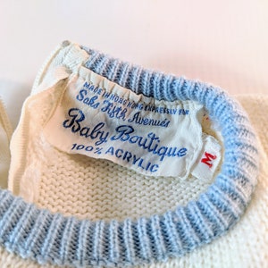May include: Close-up of a vintage baby sweater with a blue trim and a label that reads "Baby Boutique" from Saks Fifth Avenue. The sweater is cream-colored and made of 100% acrylic. The label also indicates the sweater was made in Hong Kong.