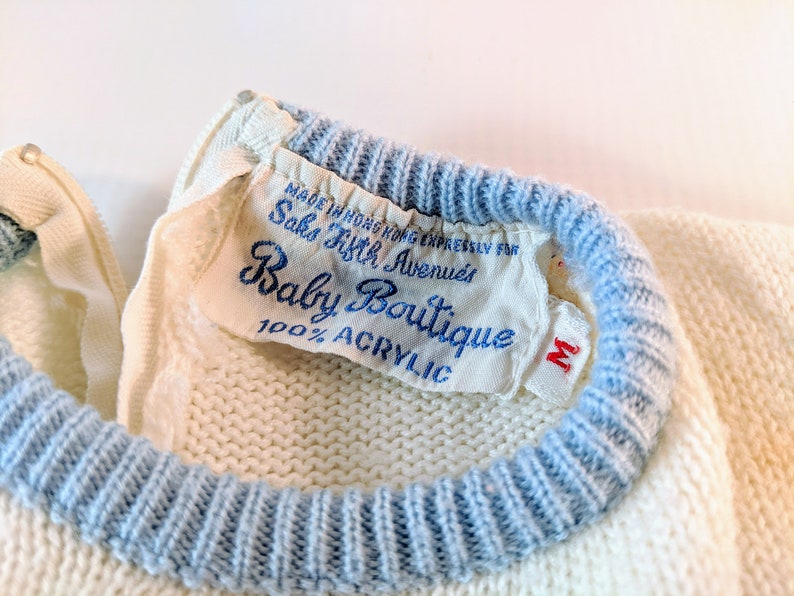 May include: Close-up of a vintage baby sweater with a blue trim and a label that reads "Baby Boutique" from Saks Fifth Avenue. The sweater is cream-colored and made of 100% acrylic. The label also indicates the sweater was made in Hong Kong.
