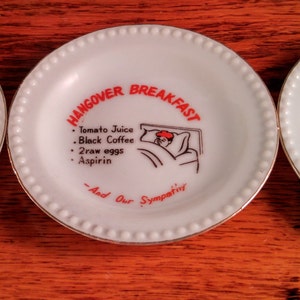 May include: Three white ceramic plates with black and red text and illustrations. The first plate says "Work, I can sit and look for hours". The second plate says "Hangover Breakfast: Tomato Juice, Black Coffee, Two eggs, Aspirin, And Our Sympathy". The third plate says "It starts when you stick your arm in his neck and ends with your arms in the sink."