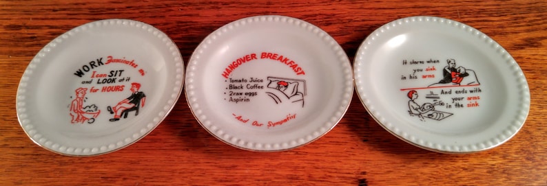 May include: Three white ceramic plates with black and red text and illustrations. The first plate says "Work, I can sit and look for hours". The second plate says "Hangover Breakfast: Tomato Juice, Black Coffee, Two eggs, Aspirin, And Our Sympathy". The third plate says "It starts when you stick your arm in his neck and ends with your arms in the sink."