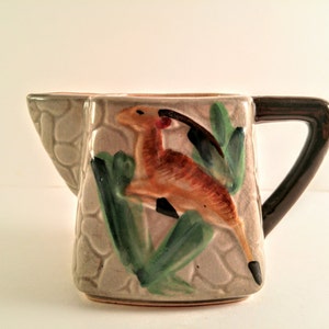May include: A ceramic creamer pitcher with a brown handle and a painted design of a deer in a jungle setting. The pitcher has a textured surface that resembles stone.