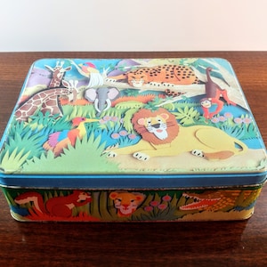 May include: A vintage tin box with a colorful jungle scene featuring a lion, giraffe, leopard, elephant, monkey, and bird. The box has a blue lid and a colorful bottom with a crocodile and squirrel.