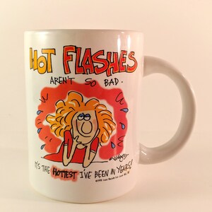 May include: White ceramic coffee mug with a cartoon illustration of a woman with red hair and a red shirt. The text on the mug reads "HOT FLASHES AREN'T SO BAD. IT'S THE HOTTEST I'VE BEEN IN YEARS!"