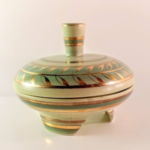 May include: A ceramic trinket box with a lid, featuring a light green base and lid with gold leaf and stripe accents. The box has three small feet and a small handle on the lid. The overall shape is round and bulbous.