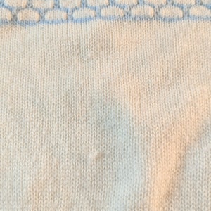 May include: Close-up of a cream-colored knit fabric with a light blue patterned border. The knit has a vertical ribbing texture. The light blue border features a repeating pattern of connected shapes.