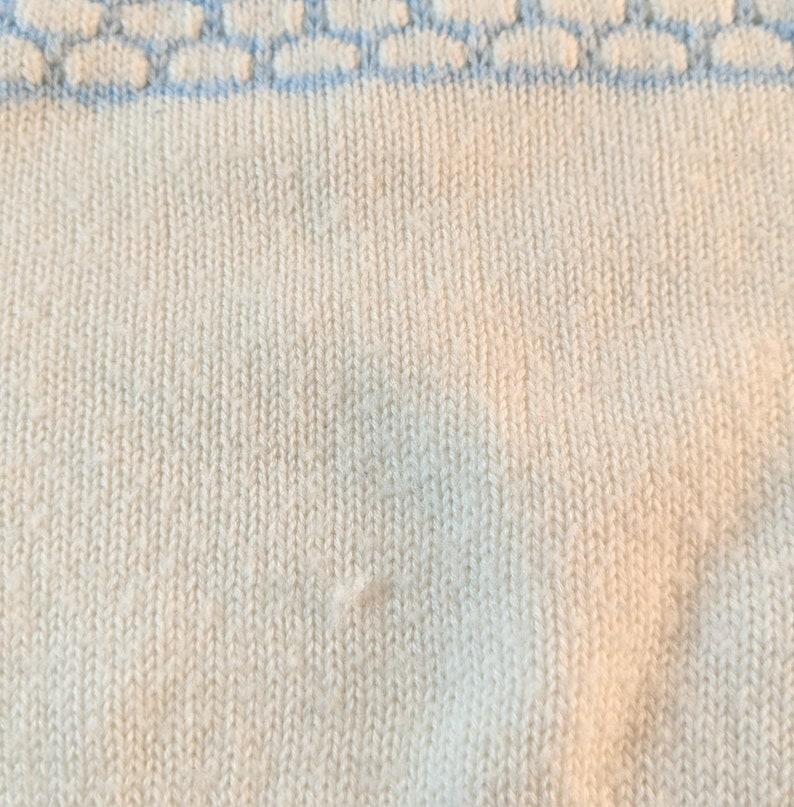 May include: Close-up of a cream-colored knit fabric with a light blue patterned border. The knit has a vertical ribbing texture. The light blue border features a repeating pattern of connected shapes.