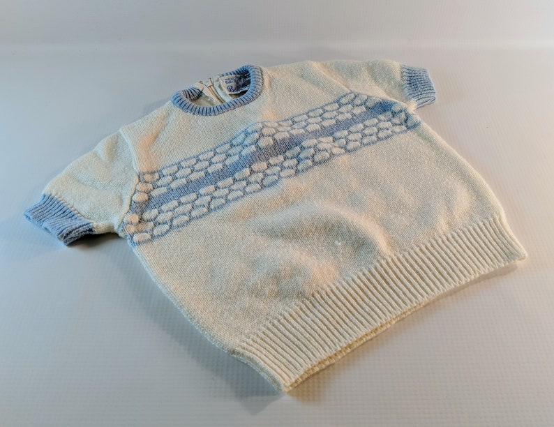 May include: Cream-colored knit sweater with short sleeves and a light blue trim. The sweater features a horizontal band of light blue textured design across the chest. The neckline and sleeve cuffs are also light blue.