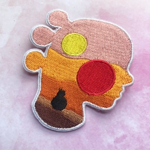 May include: Embroidered patch in the shape of a head with a cat silhouette. The patch features a pink upper section with a yellow circle, an orange and brown lower section, and a red circle. The patch is outlined in white.