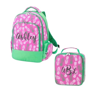 May include: A bright pink backpack and a matching lunch bag, both featuring a repeating white pineapple pattern and green trim. The backpack displays the name Ashley in black script, while the lunch bag features the monogram aBL in black.