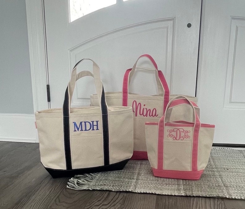 K&ouml;nnte beinhalten: Drei Stofftaschen mit schwarzen, rosa und dunkelblauen Griffen. Die Taschen sind mit den Namen "MDH", "Nina" und einem Monogramm personalisiert.