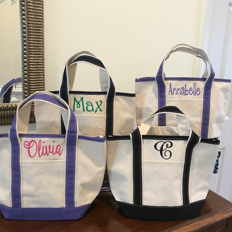 K&ouml;nnte beinhalten: Vier personalisierte Canvas-Tragetaschen mit verschiedenen farbigen Einfassungen. Die Taschen sind wei&szlig; mit schwarzen und farbigen Einfassungen. Die Taschen sind mit den Namen "Olivia", "Max", "Annabelle" und einem Monogramm "C" bestickt.