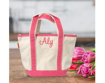 May include: A small off-white canvas tote bag featuring bright pink trim, two matching pink handles, and the name Aly embroidered in pink script on the front center. The bag has a structured base and a single exterior pocket on the front panel.