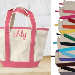 K&ouml;nnte beinhalten: Eine kleine wei&szlig;e Canvas-Tragetasche mit rosa Besatz und dem Namen "Aly" bestickt auf der Vorderseite. Die Tasche hat zwei Henkel und einen Rei&szlig;verschluss. Die Tasche wird auf einer Holzoberfl&auml;che gezeigt.
