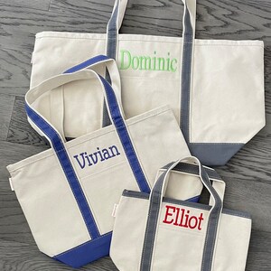 K&ouml;nnte beinhalten: Drei Stofftaschen aus Canvas mit blauem und grauem Besatz. Die gr&ouml;&szlig;te Tasche hat den Namen "Dominic" in Gr&uuml;n gestickt. Die mittlere Tasche hat den Namen "Vivian" in Blau gestickt. Die kleinste Tasche hat den Namen "Elliot" in Rot gestickt.