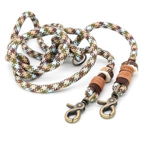 Könnte beinhalten: Eine Tarnmuster-Seilleine mit bronzefarbenen Karabinerhaken. Das Seilmuster hat Brauntöne, Grün und Blau. Die Leine hat dekorative Elemente aus Leder und Knochen, mit dem Wort "ENOS" sichtbar.