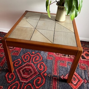 May include: A mid-century modern coffee table with a tiled top and wooden frame. The tabletop features beige tiles arranged in geometric patterns. The table is 24 inches long, 24 inches wide, and 16 inches high. A potted plant sits on the table.